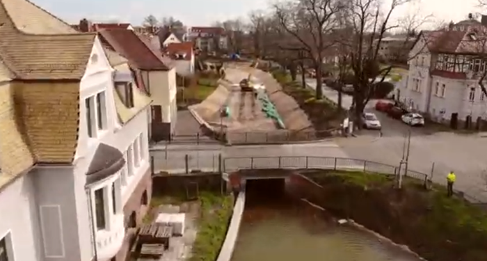 Image of moat renovation works in Delitzsch, Germany. The drone flyover image from NAUE shows housing on left, roadway on right, and in the center the moat. Half the moat is empty and with construction equipment.